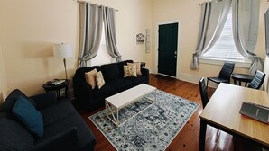 Un salon avec un canapé noir, une table basse blanche, un tapis à motifs, un ventilateur de plafond et un coin repas avec une table et des chaises.