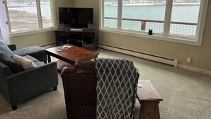 Ein Wohnzimmer mit großem Fenster mit Blick auf ein Gewässer und eine Brücke, einem Flachbildfernseher auf einem Holztisch, einem grauen Sofa, einem gemusterten Sessel und einem kleinen Holztisch.