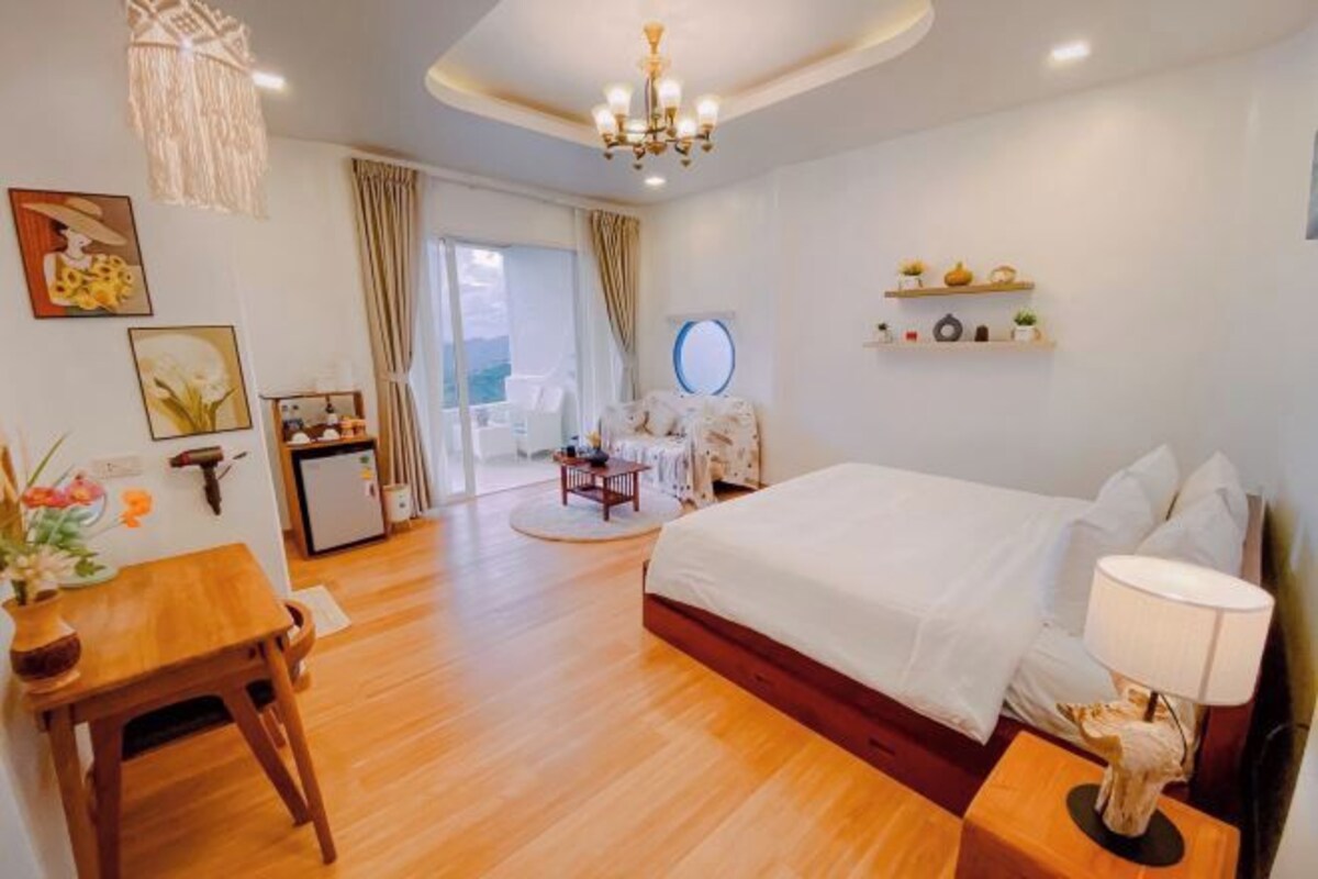 A hotel room with a large bed, a desk with a chair, a small table, a lamp, a ceiling light, a wall-mounted shelf with decorative items, and a view of the outdoors through a window.