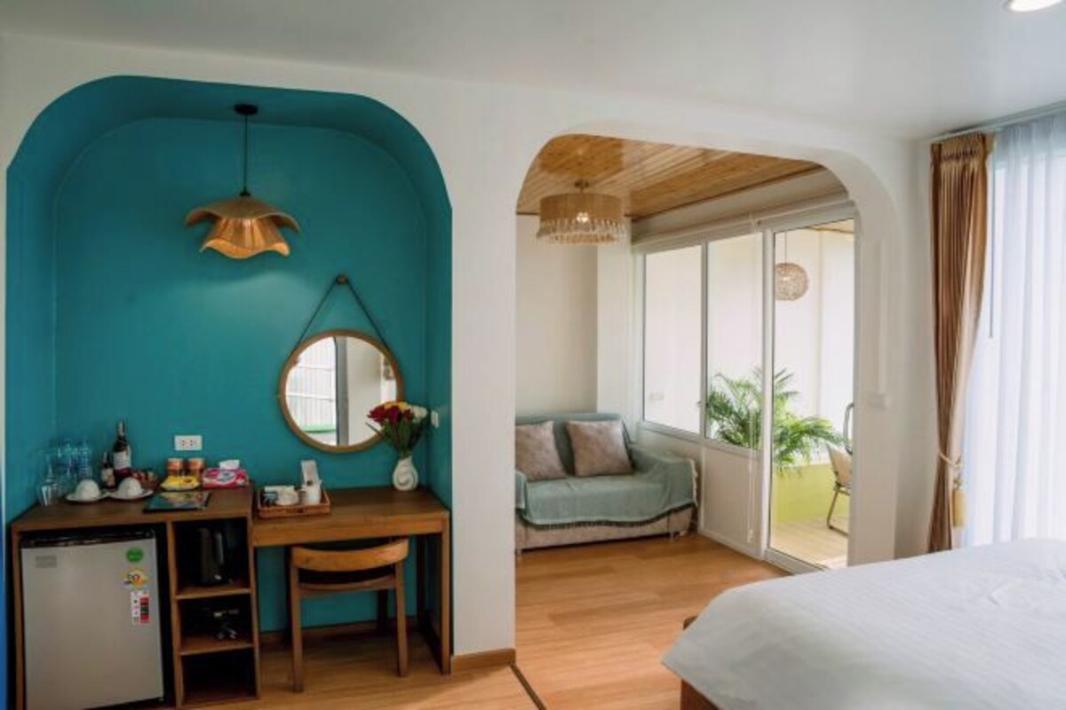 A room with a teal accent wall, a wooden desk with a mirror, a bed, and a view of a balcony with plants.