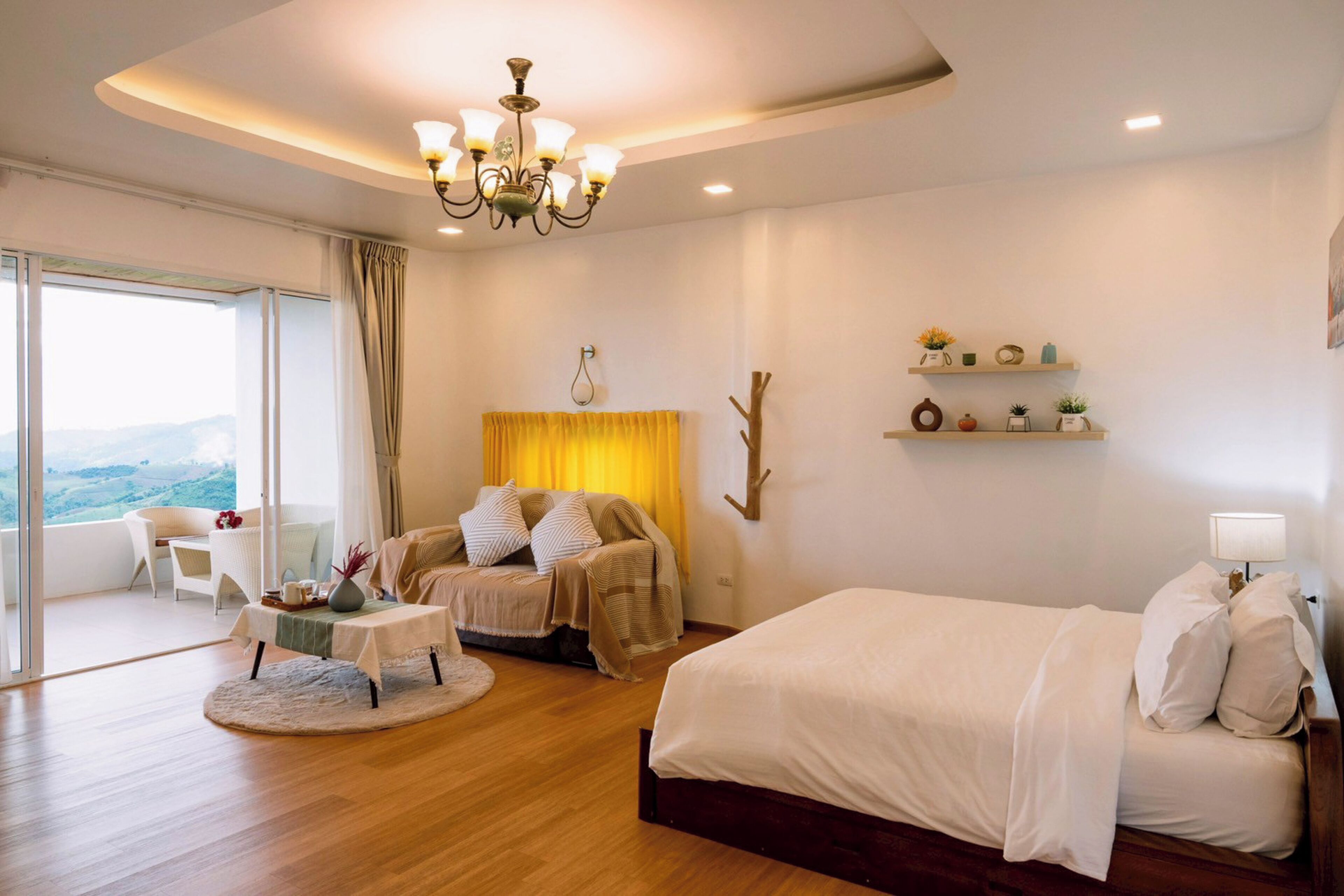 A bedroom with a bed, a sofa, a table, and a chair. There is a window with curtains and a view of the outdoors.
