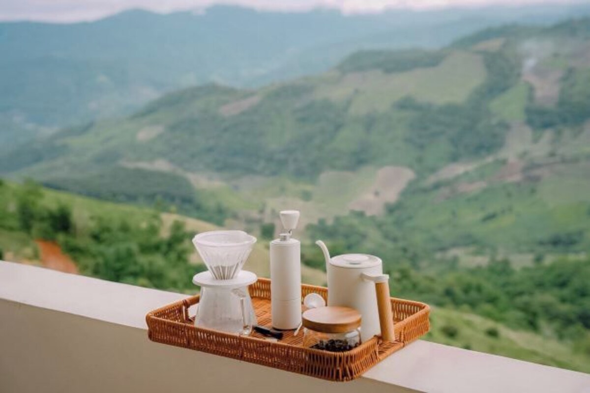 A wicker tray with a coffee maker, a kettle, and a jar on a balcony with a scenic view of green hills.