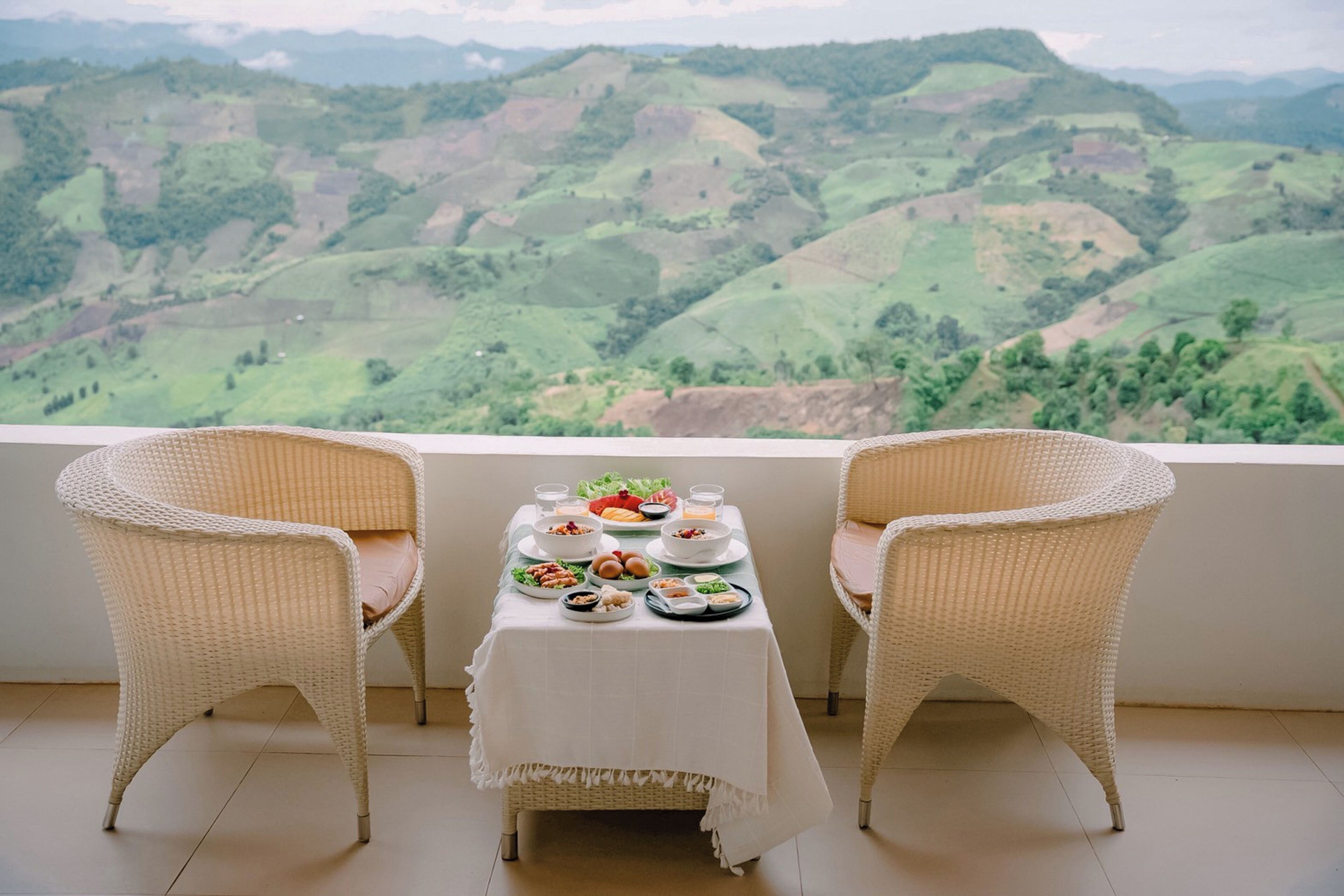 Un tavolo da pranzo con piatti di cibo e due sedie in vimini rivolte verso una vista panoramica su dolci colline.