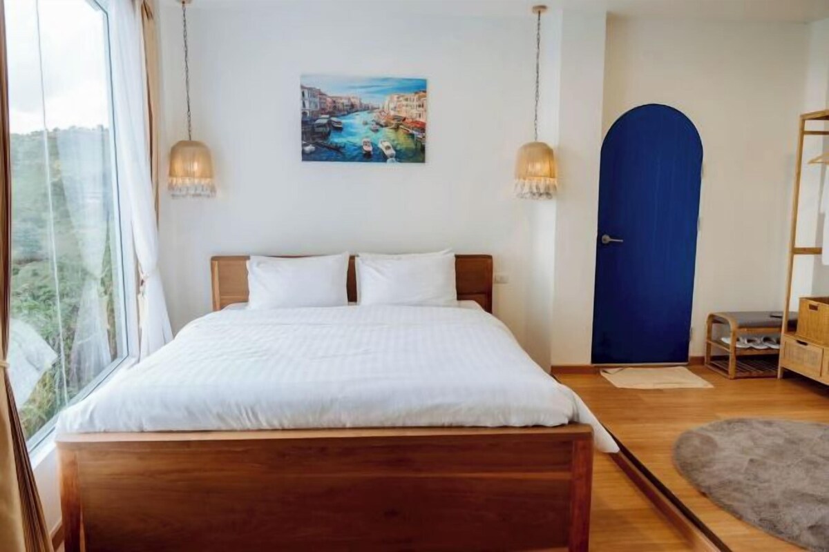 A bedroom with a wooden bed, white bedding, a blue door, and a painting of a coastal scene.