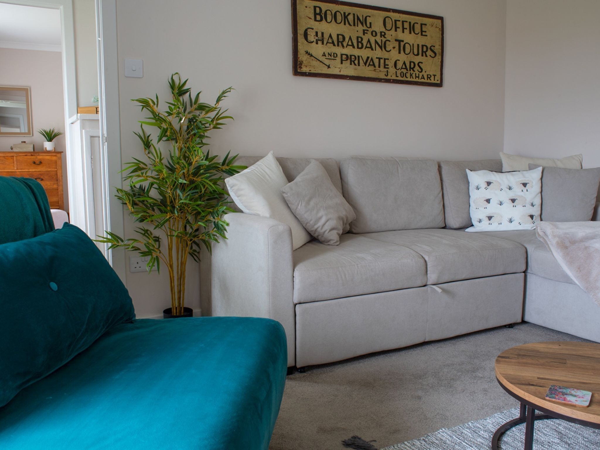 A living room with a sofa, a coffee table, and a potted plant. There is a sign on the wall that reads "Booking Office for Charabanc Tours and Private Cars."