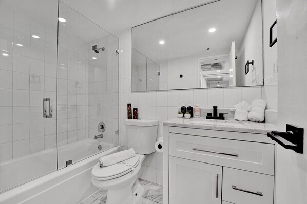 A modern bathroom with a glass-enclosed shower, a toilet, a sink with toiletries, and a vanity with drawers. - Serene Oceanfront Condo Offering Beach Service 806 (Miami Beach)