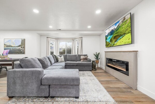 A modern living room with a grey sectional sofa, a fireplace, and a large TV mounted on the wall.