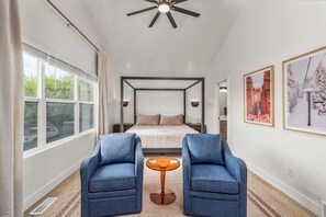 A bedroom with a large bed, two armchairs, a small table, and framed pictures on the walls.