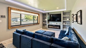 Un salon moderne avec un canapé d’angle, une cheminée, un téléviseur et une grande fenêtre donnant sur une rue et des immeubles.