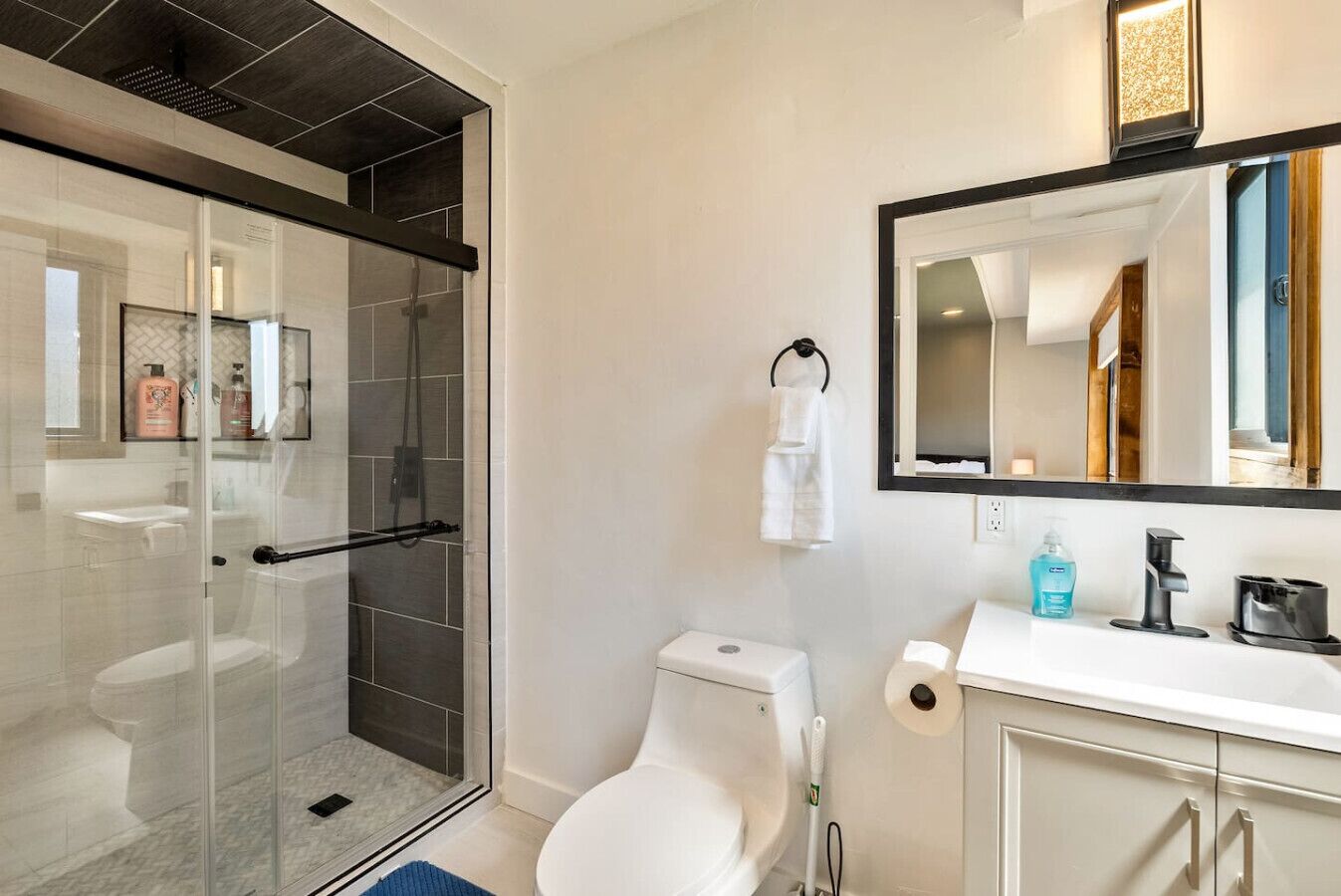 Un baño moderno con ducha envuelta en vidrio, inodoro blanco, lavamanos con espejo y toalla colgada en un gancho.