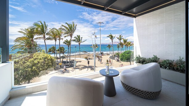 Ein Balkon mit Blick auf Strand, Palmen und Meer. Zwei weiße Stühle und ein kleiner Tisch mit einer Flasche und einem Glas.