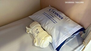 Un llit amb un coixí blanc i una tovallola estovada, una prestatgeria de fusta i una presa elèctrica a la paret.