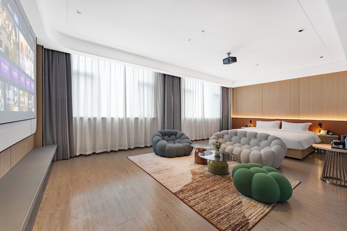 Quarto de hotel moderno com uma cama grande, uma área de estar com dois bean bags, uma pequena mesa de centro e uma janela ampla com cortinas.
