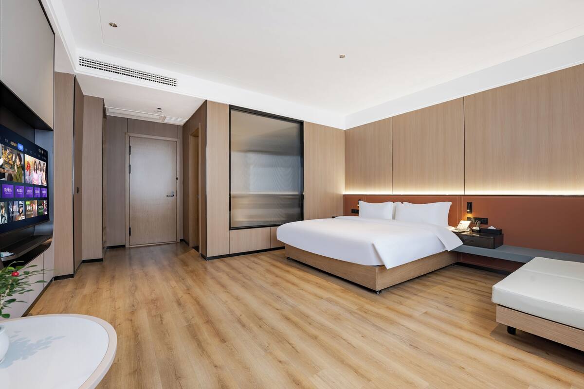 Quarto de hotel moderno com uma cama grande, uma mesinha de cabeceira, uma televisão e piso de madeira.