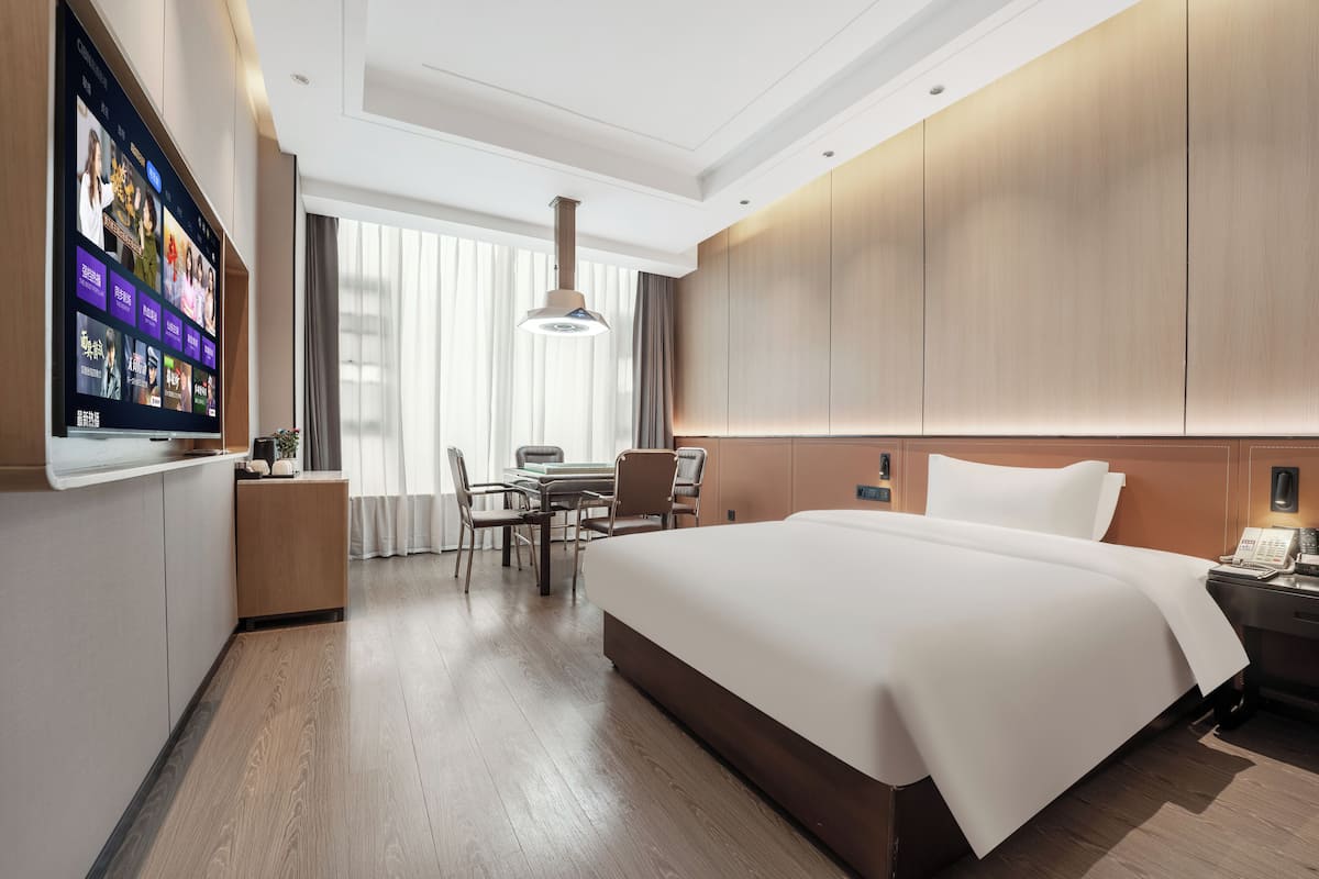 Quarto de hotel moderno com uma cama grande, uma mesa com cadeira, uma televisão na parede e uma janela com cortinas.