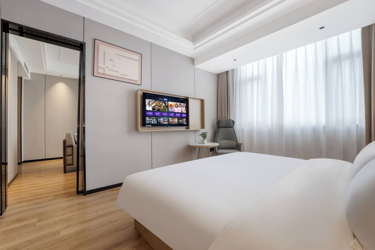 Quarto de hotel moderno com uma cama grande, uma TV fixada na parede, uma mesinha com uma planta e vista para o exterior através de cortinas leves.