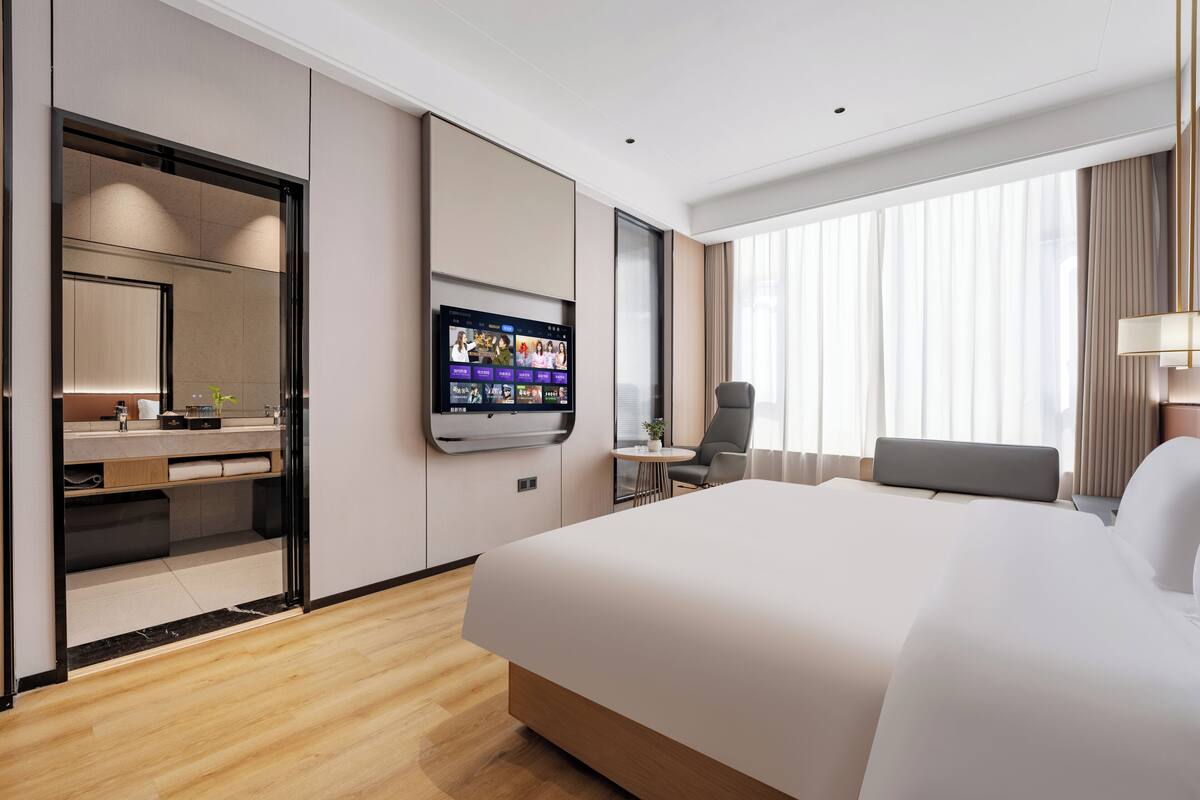 Quarto de hotel moderno com uma cama grande, televisão de tela plana na parede, uma mesa redonda pequena, uma cadeira e uma janela grande com cortinas.