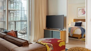 Una habitación de hotel moderna con un ventanal grande, un televisor de pantalla plana, un sofá de cuero, una cama con un edredón de colores y piso de madera.