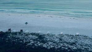A beach with white sand and calm water. There are white tents and a wooden fence along the shore.