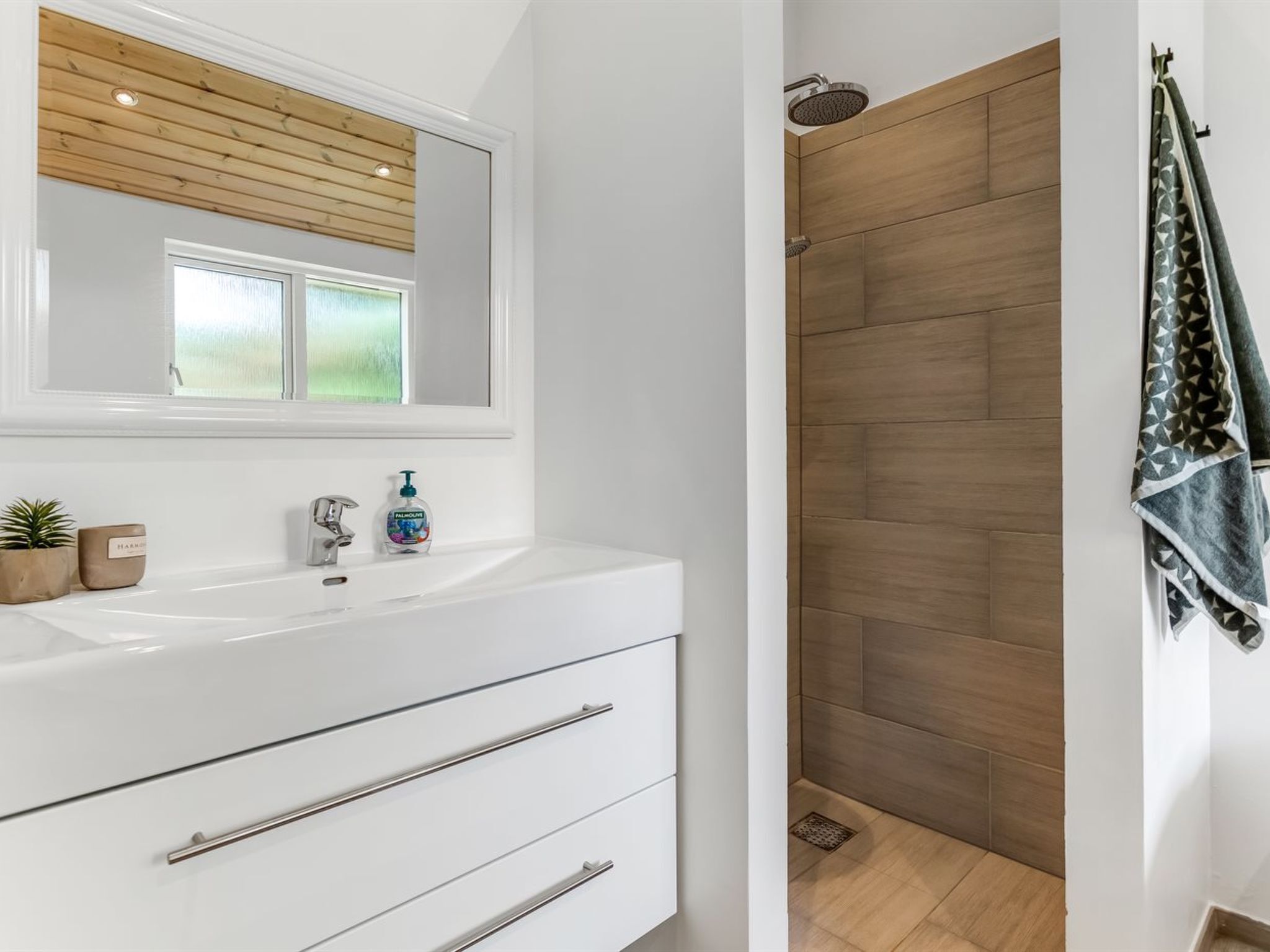 Ein modernes Badezimmer mit einem weißen Waschtisch, einem großen Spiegel und einem Duschbereich mit Holzboden.