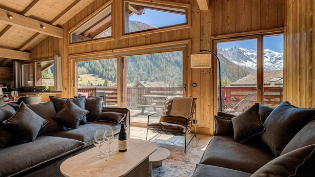 A cozy wooden interior with a sofa and armchairs, a dining table with wine glasses, and large windows offering mountain views.