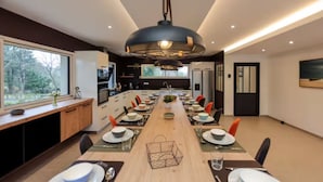 A modern dining area with a long table set for a meal, a kitchen in the background, and a large window providing a view of the outdoors.