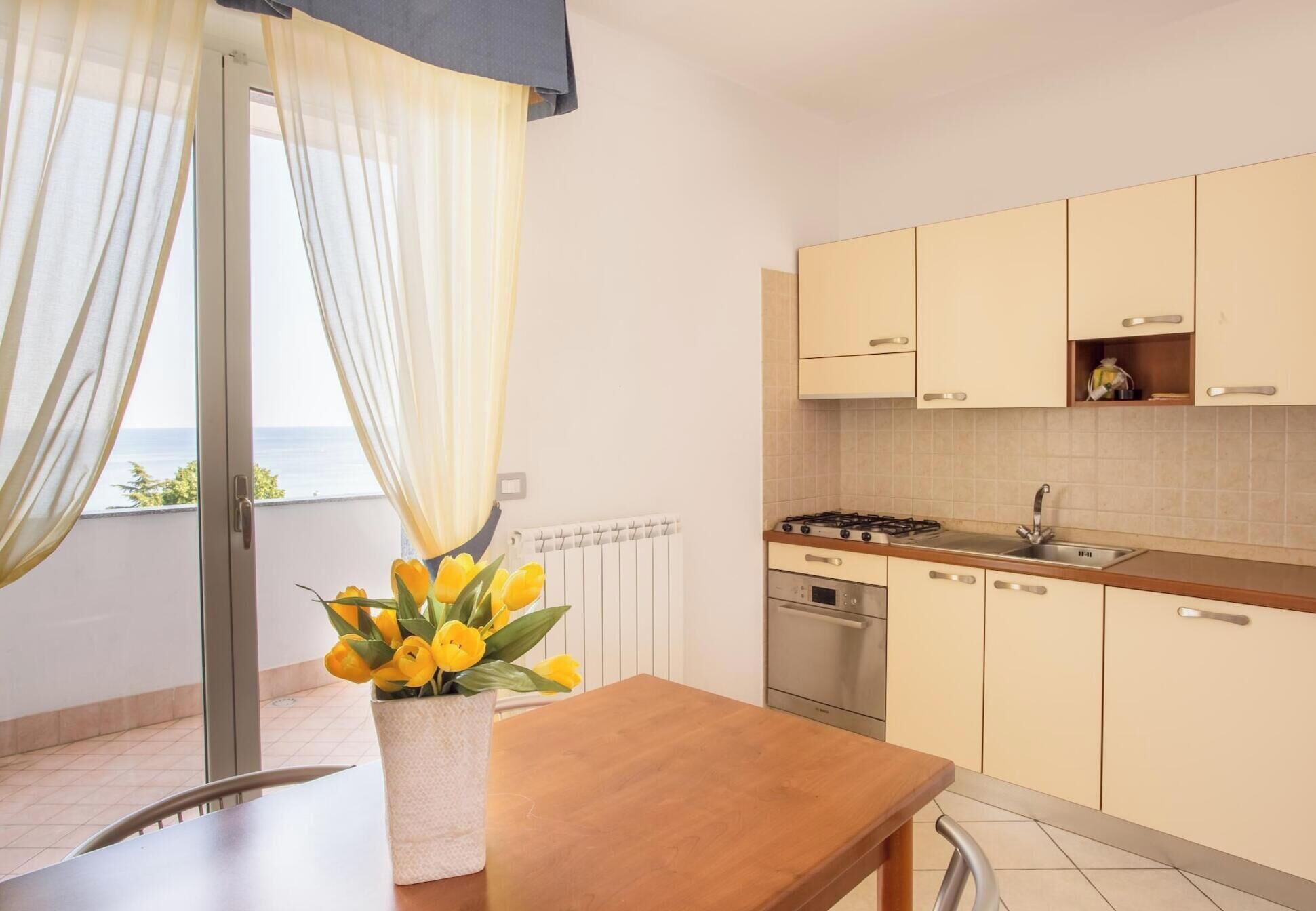 Une cuisine moderne équipée d’une cuisinière, d’un évier et de placards. Une fenêtre avec des rideaux et un vase de fleurs jaunes se trouvent sur la table.