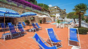 A resort pool area with blue lounge chairs and umbrellas, a stone wall with purple flowers, and a clear blue sky. - Classic Appartement in Pietra Ligure (Pietra Ligure)