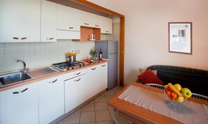 Uma cozinha compacta com armários brancos, pia de aço inoxidável e bancada de madeira. Há uma mesa de jantar com um jogo americano e uma fruteira.
