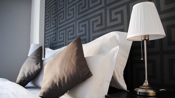 A neatly made bed with white and brown pillows, a black nightstand with a lamp, and a geometric patterned wall. - Athens Smart 3-room Private Apartment (Athens)