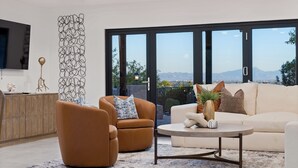 A modern living room with a sofa, armchair, and coffee table. There is a large window with a view of mountains.
