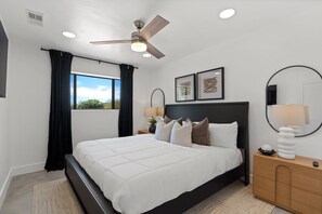 A bedroom with a bed, a ceiling fan, a nightstand with a lamp, a window with curtains, and framed artwork on the wall.