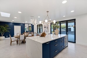 A modern kitchen with a marble island, dark cabinetry, and a dining area with chairs and a table.