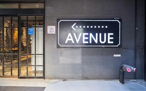 A street sign for 'AVENUE' with a left-pointing arrow, a 'BEST' sign, and a fire hydrant on the sidewalk. - AVENUE HOTEL (Osan)