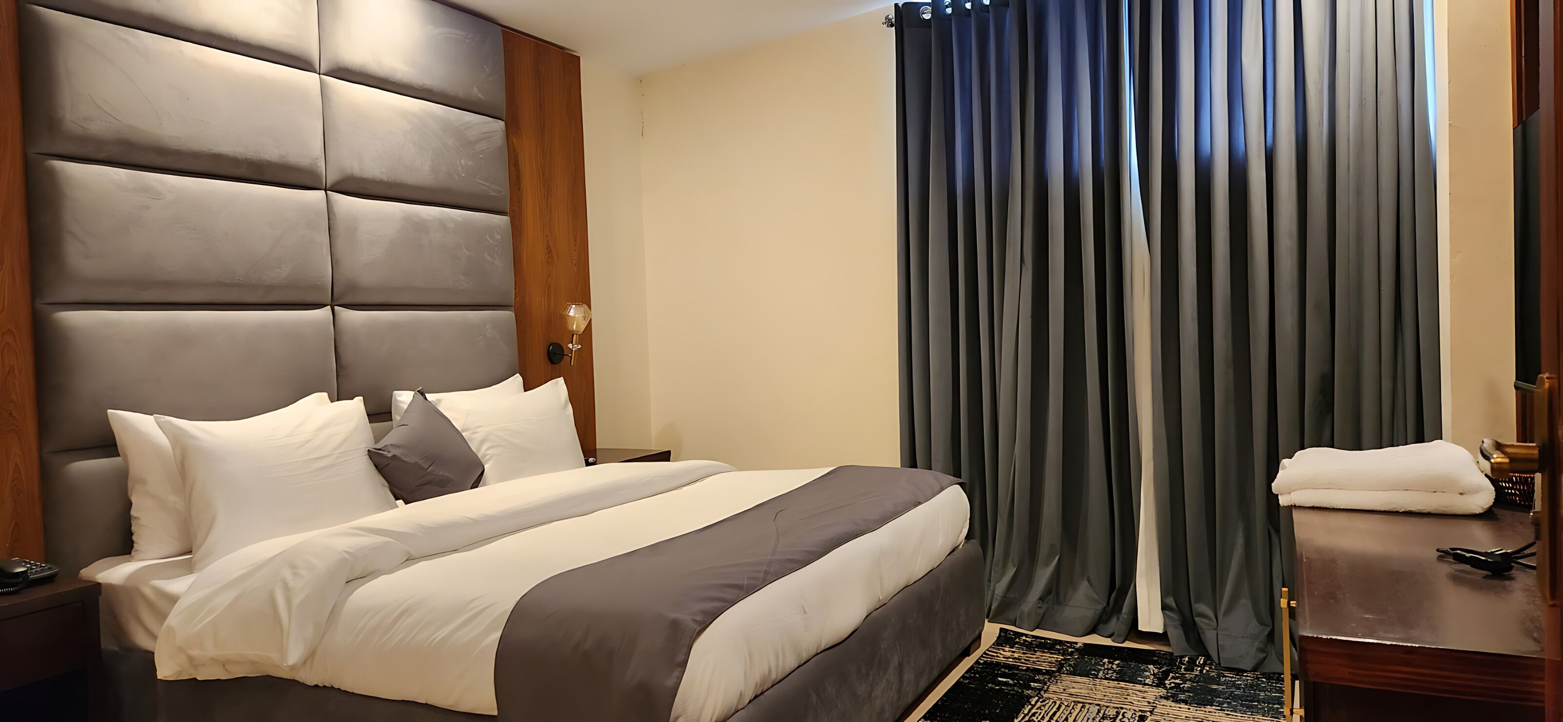 Habitación de hotel con una cama grande, un cabecero de madera, una mesita de noche con una lámpara y una ventana con cortinas oscuras.