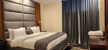 Habitación de hotel con una cama grande, un cabecero de madera, una mesita de noche con una lámpara y una ventana con cortinas oscuras.