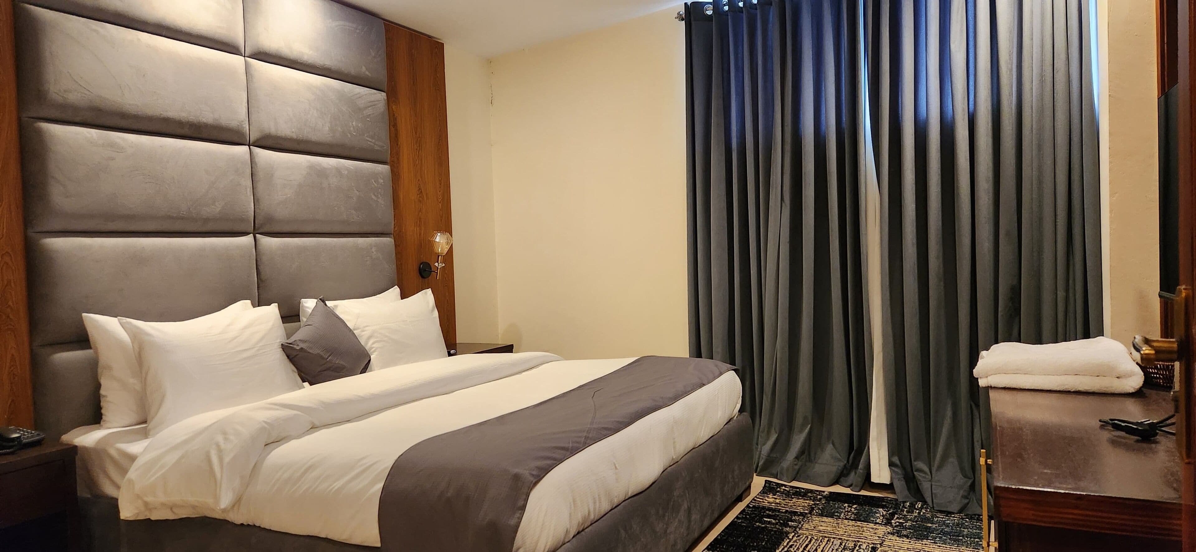A hotel room with a large bed, a wooden headboard, a bedside table with a lamp, and a window with dark curtains.
