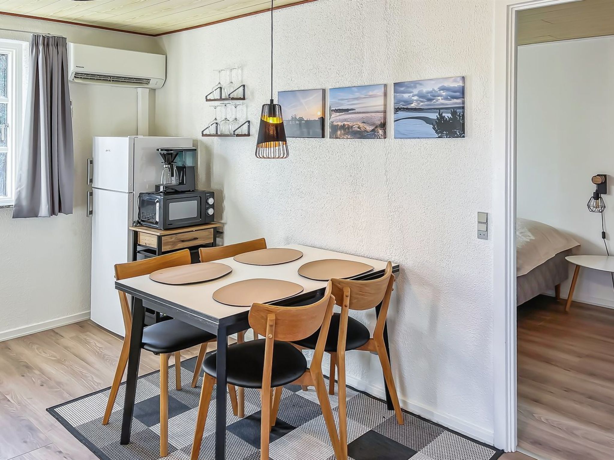 Eine kompakte Küche mit Kühlschrank, Mikrowelle und Kaffeemaschine. Ein Esstisch mit vier Stühlen steht dort, darüber hängt eine Pendelleuchte. Die Wände sind mit drei gerahmten Bildern dekoriert.