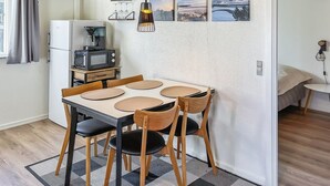 Une cuisine compacte équipée d’un réfrigérateur, d’un four à micro-ondes et d’une machine à café. On y trouve une table à manger avec quatre chaises et un luminaire suspendu. Les murs sont décorés de trois tableaux encadrés.