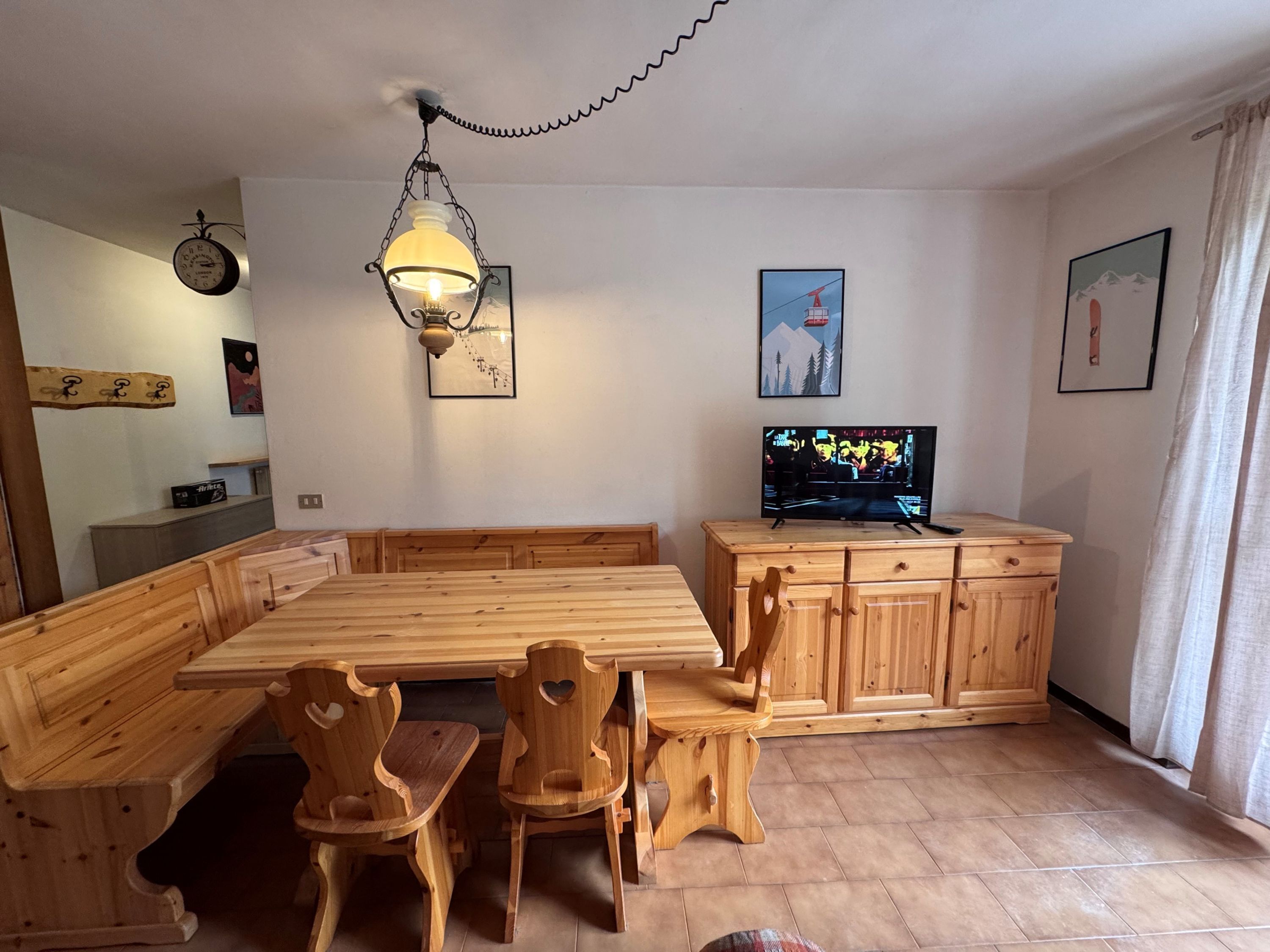 Een eetgedeelte met een houten tafel en stoelen, een televisie op een houten kast, en een hangende lamp.