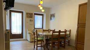 A dining area with a wooden table and chairs, a radiator, and two windows with curtains. - 020 Trilocale Pinzolo (Pinzolo)
