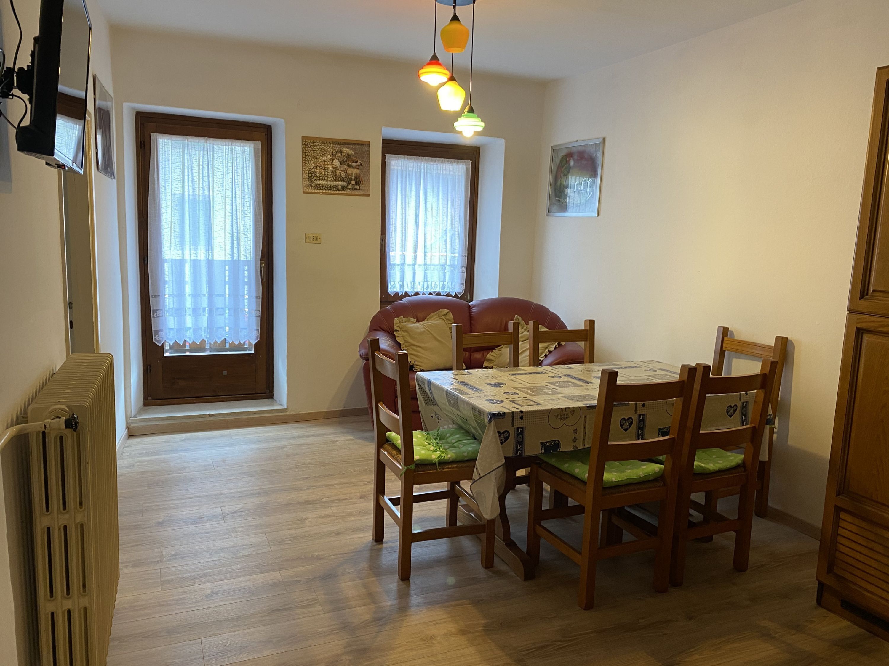 A dining area with a table set for four, surrounded by wooden chairs with green cushions. There are two windows with curtains, a radiator, and a ceiling light fixture.