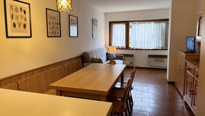 Ein gemütlicher Essbereich mit Holztisch und -stühlen, einer Bank und einem Sofa. An der Wand befinden sich gerahmte Pflanzenbilder, eine Pendelleuchte und ein Fenster mit Vorhängen.