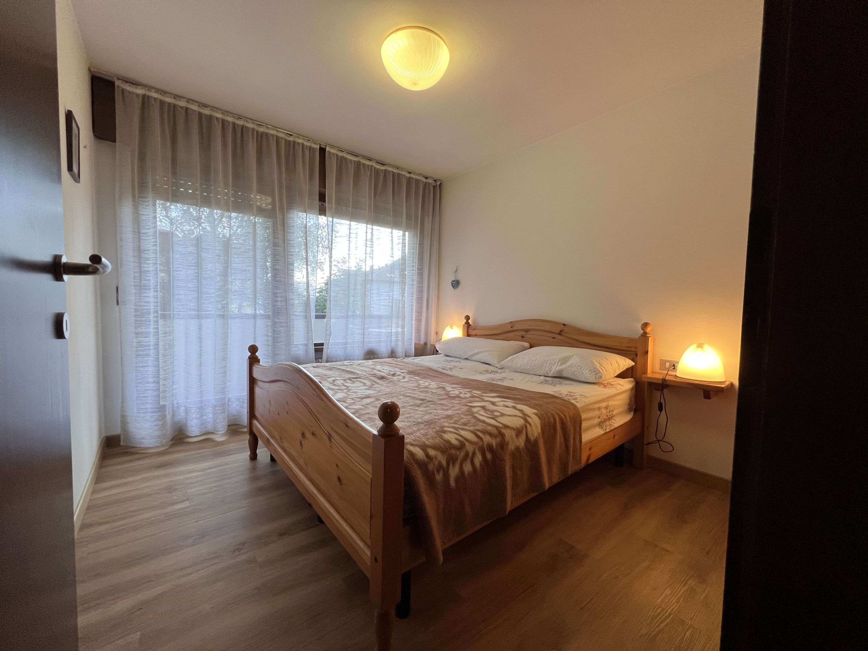 A bedroom with a wooden bed, white curtains, a bedside lamp, and a ceiling light.