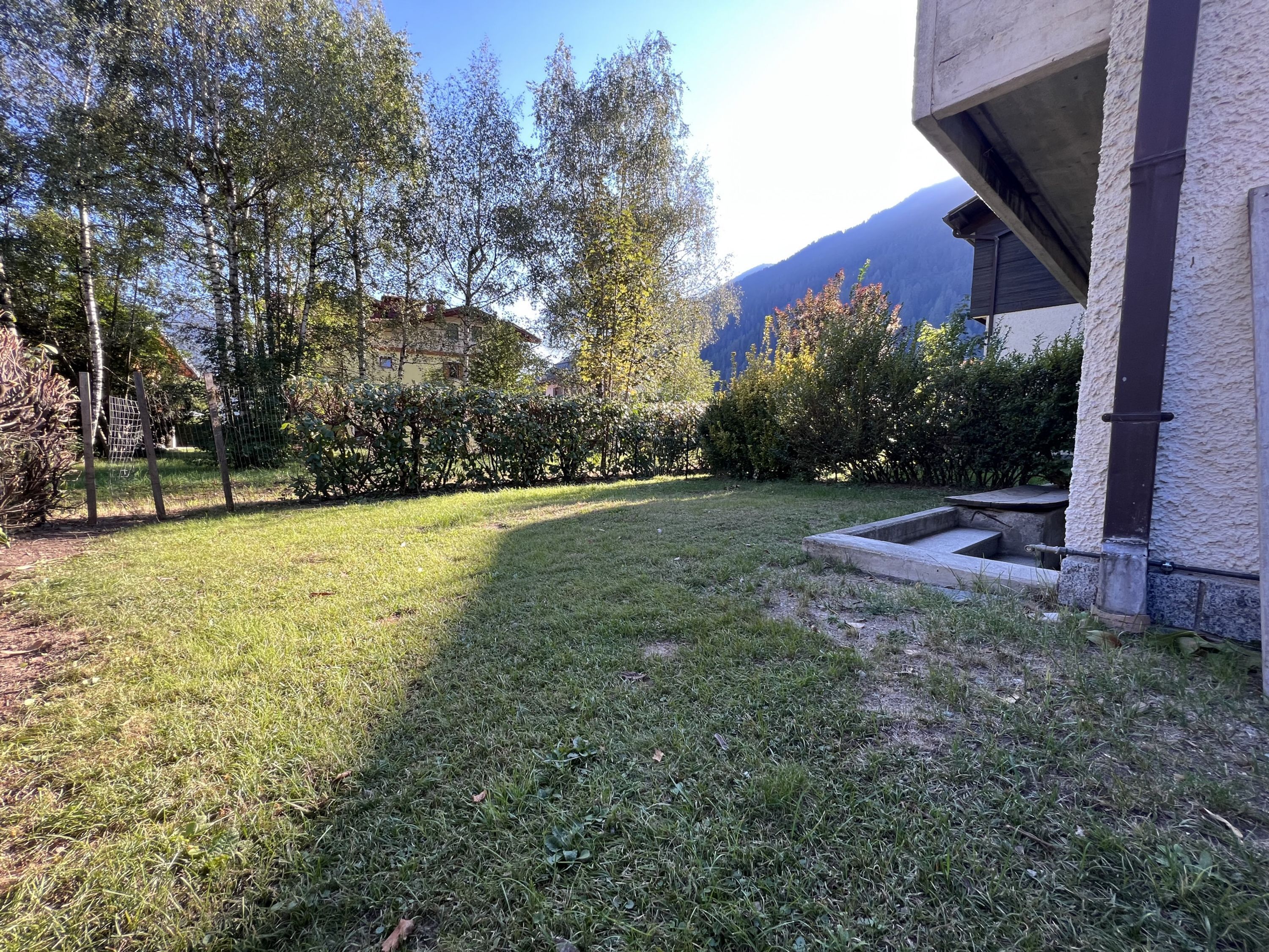 Un jardín césped cuidado con un macetero de piedra, cerca de madera y un edificio con pared exterior texturizada.
