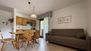 A spacious living area with a brown sofa, wooden dining table, and chairs. A kitchen with wooden cabinets and a microwave is visible in the background. - 091 Cond Gaiolin 23 (Giustino)