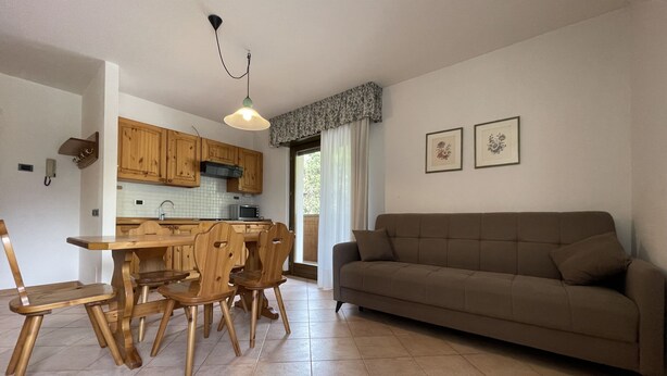 A spacious living area with a brown sofa, wooden dining table, and chairs. A kitchen with wooden cabinets and a microwave is visible in the background. - 091 Cond Gaiolin 23 (Giustino)