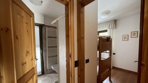 A hallway with wooden doors, a white tiled floor, and a view into a room with a bed and a window with curtains. - 091 Cond Gaiolin 23 (Giustino)