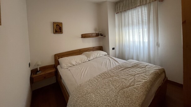 A bedroom with a wooden bed, a nightstand, a wall-mounted shelf, and a window with sheer curtains. - 091 Cond Gaiolin 23 (Giustino)