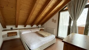 A bedroom with a wooden ceiling, a bed with a mattress and pillows, a bedside table, a radiator, and a window with curtains. - 092 Cond Gaiolin 27 (Giustino)